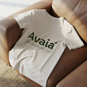 Op de afbeelding: Een wit t-shirt met het woord "Avaia" in donkergroen, samen met "PSD MOCKUPS JPG". Het t-shirt is gedrapeerd over een bruine leren fauteuil, wat een casual en ontspannen uitstraling creëert. Het t-shirt is een mockup.