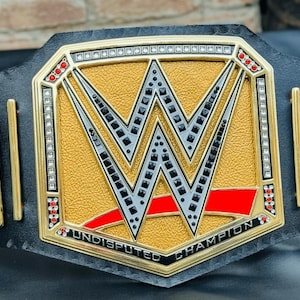 May include: A gold and black championship belt with a large, gold, octagonal center plate featuring a black and white "W" logo and the text "Undisputed Champion".