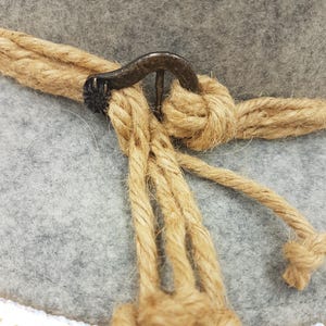 May include: Close-up of a gray felt hat with a natural rope band and decorative rope detail. The hat features a dark metal clasp and a white crocheted brim. The rope is knotted around the hat.