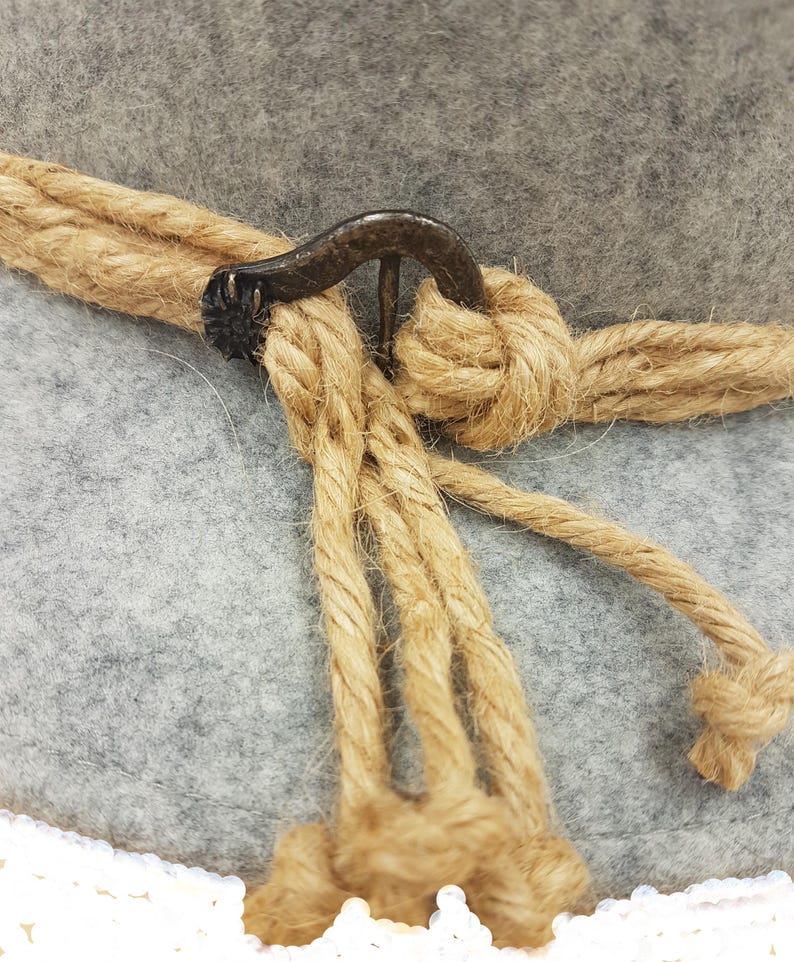 May include: Close-up of a gray felt hat with a natural rope band and decorative rope detail. The hat features a dark metal clasp and a white crocheted brim. The rope is knotted around the hat.