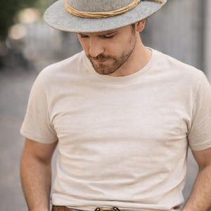 May include: A gray fedora-style hat with a tan rope band and a decorative knot. The hat is worn with a light-colored t-shirt and a brown leather belt with a decorative buckle. The buckle has a heraldic design.