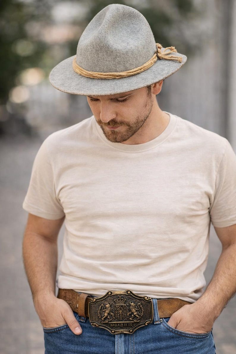 May include: A gray fedora-style hat with a tan rope band and a decorative knot. The hat is worn with a light-colored t-shirt and a brown leather belt with a decorative buckle. The buckle has a heraldic design.