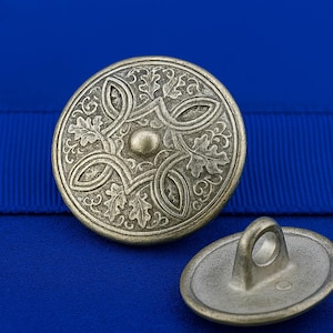Puede incluir: Primer plano de dos botones metálicos de estilo antiguo. El botón más grande presenta un intrincado diseño en relieve con un botón central y un patrón de hojas y formas entrelazadas. El botón más pequeño tiene un lazo para coser. Los botones están sobre un fondo azul.