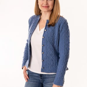 Könnte beinhalten: Eine Frau trägt einen blauen Strickcardigan mit silbernen Knöpfen über einem weißen V-Ausschnitt-Shirt und blauen Jeans. Der Cardigan hat ein strukturiertes Muster und einen runden Ausschnitt. Sie lächelt vor einem weißen Hintergrund.