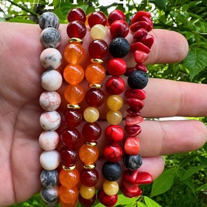 May include: Five beaded bracelets in various colours and textures. The bracelets feature a mix of round and irregular beads in shades of red, orange, black, grey, and white. Some bracelets have gold-coloured spacers.