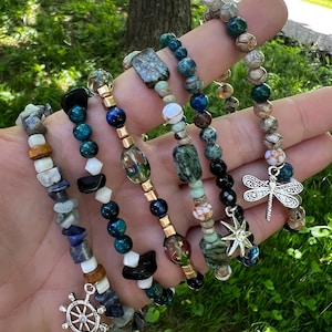 May include: Six beaded bracelets in various colours are displayed. The bracelets feature natural stone beads, gold accents, and charms, including a ship's wheel, a star, and a dragonfly. The bracelets are displayed against a green background.