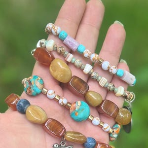 May include: Several beaded bracelets with a mix of natural stone beads in brown, yellow, blue, and turquoise. The bracelets have gold-coloured accents and a small bird charm. The beads are of different sizes and shapes.