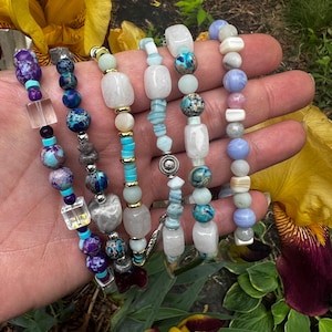 May include: Several beaded bracelets in shades of turquoise, blue, purple, and white. The bracelets incorporate stone and glass beads, some with gold detailing. The bracelets are shown against a natural backdrop with flowers.