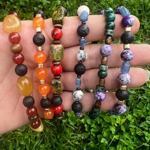May include: Six beaded bracelets featuring a variety of stone and lava rock beads in shades of yellow, orange, red, green, blue, and purple. The bracelets are displayed against a backdrop of green grass.