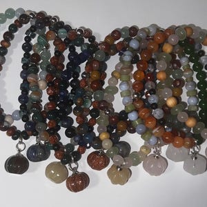 May include: A selection of beaded bracelets in shades of green, brown, blue, and orange. Each bracelet has different beads and a small pumpkin-shaped charm. The bracelets are displayed on a white surface.