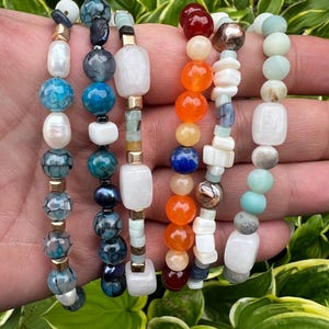 May include: Six beaded bracelets with a variety of colourful beads, including blue, white, orange, and green. The bracelets feature gold-coloured spacers and a mix of round and rectangular beads. The bracelets are displayed against a backdrop of green foliage.