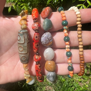 May include: Five beaded bracelets with diverse designs and materials. One bracelet features a large, patterned bead, while others showcase colourful stones and wooden beads. The bracelets are held up against a background of green foliage.