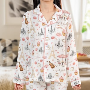 May include: White pajama set with a repeating pattern of the Eiffel Tower, chandeliers, musical instruments, and cafe scenes in pink and beige. The set includes a button-down top and pants, trimmed with pink piping.