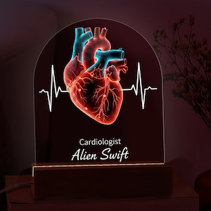 Może przedstawiać: Dekoracyjna lampka nocna z wystylizowanym ludzkim sercem z niebieskimi i czerwonymi akcentami oraz białą linią EKG. Tekst "Kardiolog Alien Swift" jest wyświetlany pod obrazem serca. Światło spoczywa na drewnianej podstawie.