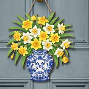 以下が含まれることがあります： 黄色と白の水仙の花束が描かれた木製のドアハンガー。青と白のベースに「M」の文字が入っています。ロープが付いた金のドアノッカーから吊り下げられています。