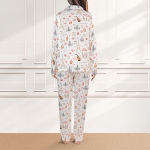 May include: White pajama set with a repeating pattern of the Eiffel Tower, chandeliers, musical instruments, and cafe scenes in pink and beige. The long-sleeved top and pants are visible.