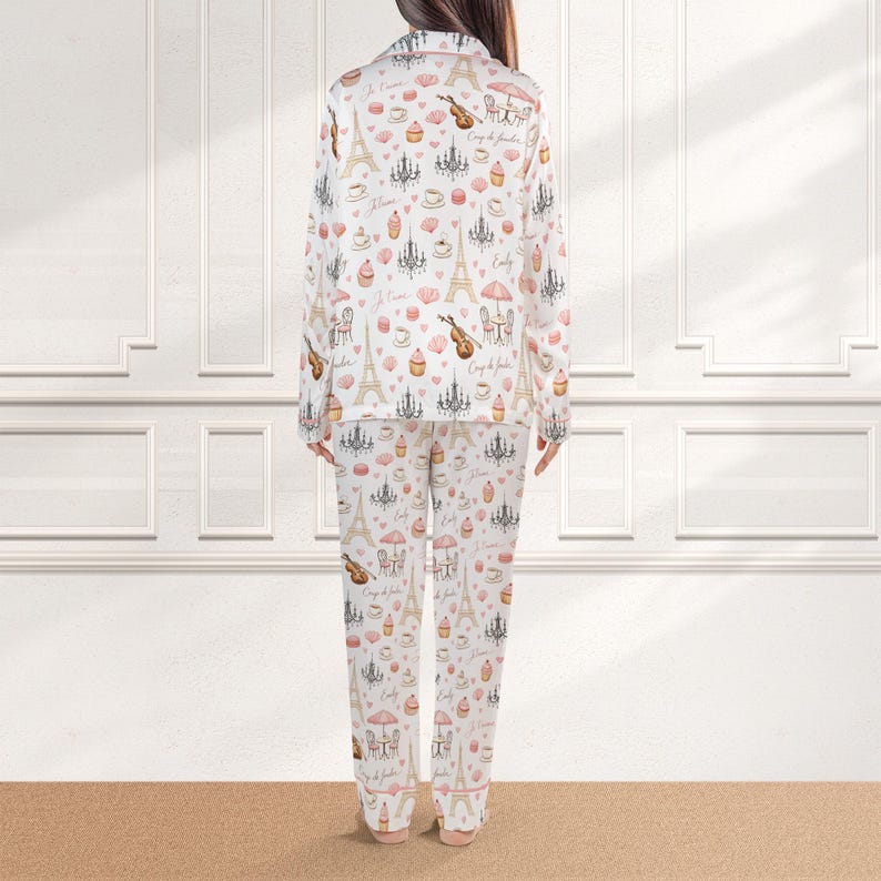 May include: White pajama set with a repeating pattern of the Eiffel Tower, chandeliers, musical instruments, and cafe scenes in pink and beige. The long-sleeved top and pants are visible.
