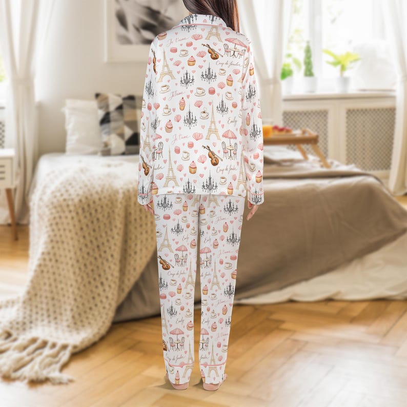May include: White pajama set with a repeating pattern of Parisian icons, including the Eiffel Tower, chandeliers, and cafe scenes in pink and brown. The long-sleeved top and pants are visible.