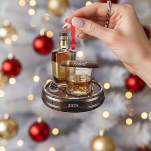 May include: A whiskey-themed Christmas ornament featuring a bottle of whiskey, a glass of whiskey with ice cubes, and a cigar with the name "Robert". The ornament is set on a wooden barrel base with the year "2025" and a red ribbon for hanging.