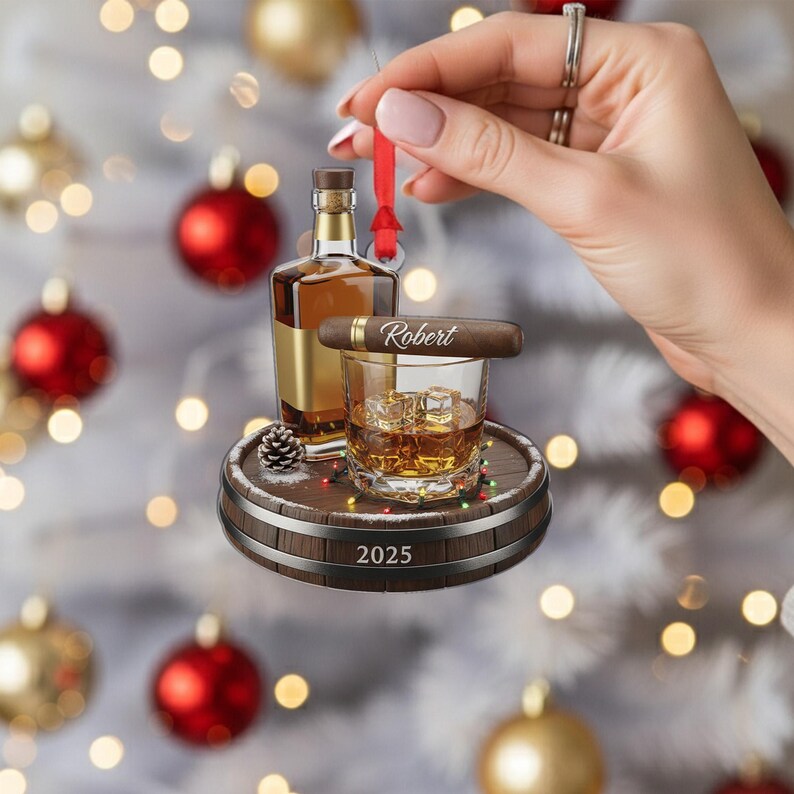 May include: A whiskey-themed Christmas ornament featuring a bottle of whiskey, a glass of whiskey with ice cubes, and a cigar with the name "Robert". The ornament is set on a wooden barrel base with the year "2025" and a red ribbon for hanging.