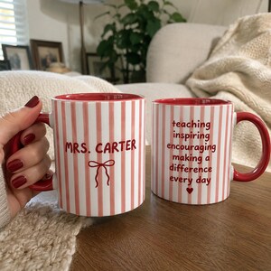 Puede incluir: Dos tazas de cerámica blanca con rayas verticales rosas y texto en rojo. Una taza presenta el texto "MRS. CARTER" sobre un gráfico de lazo. La otra taza muestra el texto "teaching inspiring encouraging making a difference every day" con un corazón.