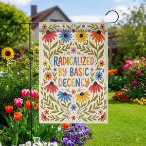 Op de afbeelding: Een tuinvaandel met de tekst "RADICALIZED BY BASIC DECENCY" in kleurrijke letters. De vlag heeft een bloemenontwerp met rode, blauwe, gele en oranje bloemen en groene bladeren. De vlag wordt getoond in een tuin met bloemen en groen.