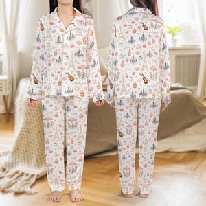 May include: White pajama set with a pink trim and a repeating pattern of Parisian icons, including the Eiffel Tower, chandeliers, and musical instruments. The set includes a button-down top and matching pants. The text "Emily" is printed on the pants.