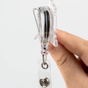 May include: A clear plastic retractable badge holder with a silver clip and a silver button. The holder is attached to a clear plastic strap with a silver ring.