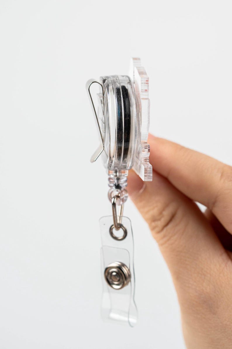 May include: A clear plastic retractable badge holder with a silver clip and a silver button. The holder is attached to a clear plastic strap with a silver ring.