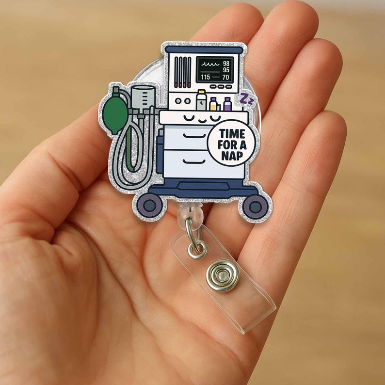May include: A silver glitter badge reel with a cartoon illustration of a medical breathing machine. The machine has a screen that reads "98, 95, 115, 70" and a small bed with a "Zzz" above it. The text "Time for a Nap" is on a blue circle.