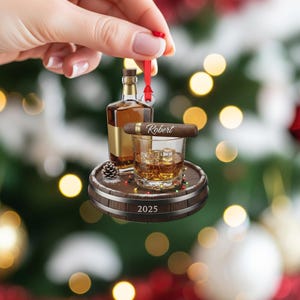 May include: A decorative ornament featuring a miniature whiskey bottle, a glass of whiskey with ice cubes, and a cigar with the name "Robert". The items are arranged on a wooden barrel-shaped base with the year "2025" and a red ribbon for hanging.