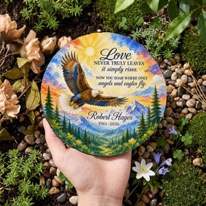 May include: A round memorial plaque featuring a colorful illustration of a bald eagle soaring over a mountain landscape with pine trees and a sunburst. The text reads: Love NEVER TRULY LEAVES it simply rises. NOW YOU SOAR WHERE ONLY angels and eagles fly. Robert Hayes 1961-2026.