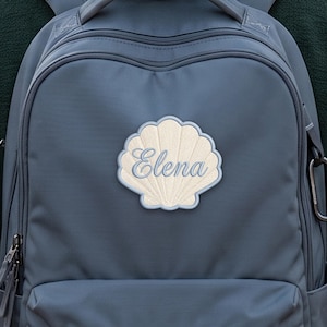 Peut inclure: Un sac à dos bleu ardoise orné d'un écusson brodé en forme de coquillage au centre. L'écusson est de couleur crème avec une bordure bleu clair et le prénom Elena est brodé en lettres cursives bleu clair au milieu du coquillage.