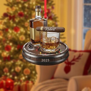 May include: A Christmas ornament featuring a whiskey bottle, a glass of whiskey with ice cubes, and a cigar with the name "Robert" on it. The ornament is set on a miniature barrel with the year "2025" and a pinecone.