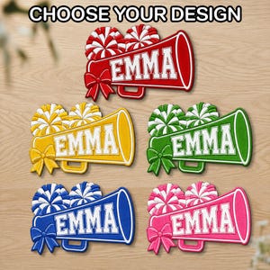 May include: Five cheerleading megaphone cutouts with pom-poms and bows, each featuring the name EMMA in white block letters. The designs are red, yellow, green, blue, and pink. The text CHOOSE YOUR DESIGN is displayed at the top of the image.
