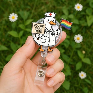 Op de afbeelding: Een badge met een witte eend in een verpleegstersuniform, met een regenboogvlag en een bord met de tekst "YOU'RE SAFE HERE". De badge heeft een transparante plastic band en een metalen clip. De achtergrond is groen met witte bloemen.