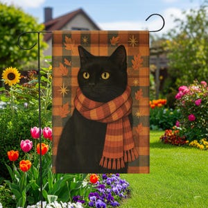 Puede incluir: Una bandera de jardín con un gato negro con una bufanda naranja y marrón. El gato está sobre un fondo a cuadros con hojas de otoño y estrellas. La bandera se muestra en un jardín con flores.