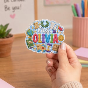 May include: A scalloped, light blue gingham embroidered patch featuring the text Ms. OLIVIA in colorful letters. The design includes icons of a globe, a history book, a scroll, a laurel wreath, a column, a quill, a flag, a map, and an hourglass.