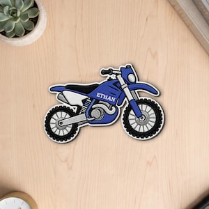 Puede incluir: Un parche bordado en azul y blanco con forma de motocicleta de cross. El cuerpo de la moto presenta el nombre ETHAN en letras mayúsculas blancas. El diseño incluye neumáticos negros con radios plateados, un asiento negro y paneles laterales blancos.