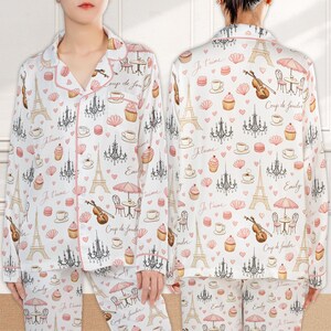 May include: White pajama set with a pink trim, featuring a repeating pattern of the Eiffel Tower, chandeliers, cupcakes, and coffee cups. The set includes a button-down top and matching pants. The words "Je t'aime", "Coup de foudre", and "Emily" are also printed on the pajamas.