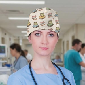May include: A beige surgical scrub cap featuring a repeating pattern of cartoon frogs driving cars in yellow, blue, and green. The text "BEEP BEEP BITCH" is also printed on the cap. The cap is worn by a person in a blue scrub top.