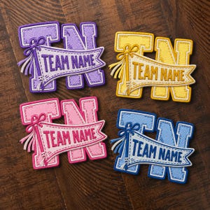 Op de afbeelding: Vier geborduurde patches in paars, geel, roze en blauw. Elke patch heeft een grote blokletter N achter een wimpel met een kwastje en de tekst TEAM NAME. Het stiksel is zichtbaar op elk stuk, wat het gelaagde ontwerp goed laat zien.