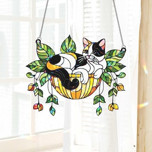 May include: Stained glass cat suncatcher. A calico cat rests in a yellow and orange basket with green leaves and hanging flowers. The cat is black, white, and orange. The suncatcher hangs from a silver chain.