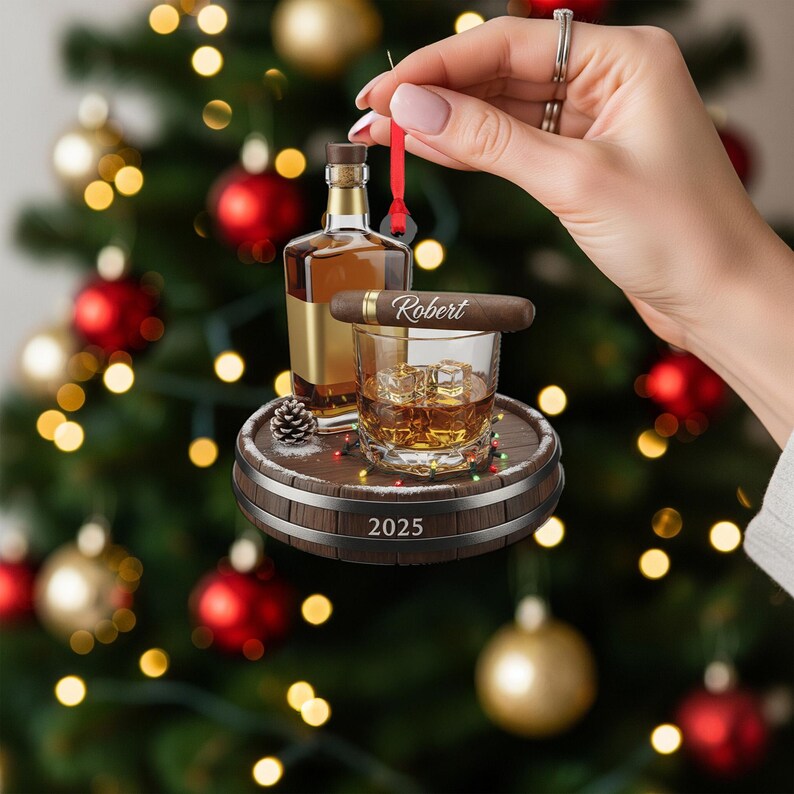 May include: A Christmas ornament featuring a miniature whiskey bottle, a cigar with the name "Robert", and a glass of whiskey with ice cubes. The ornament is set on a wooden barrel base with the year "2025" and festive lights.