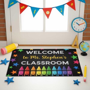 以下が含まれることがあります： 「WELCOME to Ms. Stephen's CLASSROOM」の文字とカラフルなクレヨンが描かれた黒い教室のドアマット。マットは星と両側の鉛筆で飾られています。時計、地球儀、バナーも表示されています。