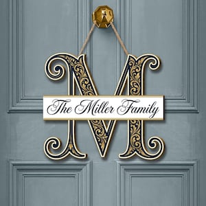 Puede incluir: Un letrero de puerta de madera con una gran letra "M" adornada en azul marino y dorado, con las palabras "The Miller Family" en una pancarta blanca. El letrero cuelga de un aldaba dorada en una puerta azul.