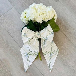 May include: A bouquet of white hydrangeas tied with a large bow. The bow features a white background with light blue stripes and a delicate pattern of green swirls and blue accents. The flowers and bow are set against a light gray wood-grain surface.