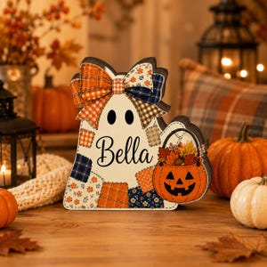 May include: A layered wooden ghost decoration featuring a patchwork pattern of orange, navy, and white fabrics. The ghost has a large bow on top, the name Bella in black script, and holds an orange jack-o'-lantern bucket filled with autumn leaves.