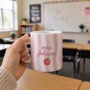 May include: White ceramic mug with pink vertical stripes, featuring the text "mrs. Johnson" and a red apple graphic. The mug is held in a classroom setting, suggesting a teacher-themed product.