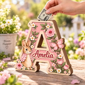 Può includere: Un salvadanaio in legno a forma di lettera A con sfondo rosa, decorato con fiori bianchi e rosa, foglie verdi, una farfalla rosa e un coniglio marrone. Il nome Amelia è scritto in rosso e vi è inserita una banconota da un dollaro.
